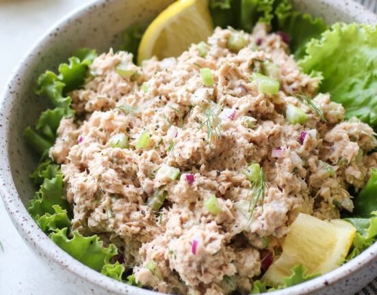 A speckled white bowl holding fresh salmon salad that is laying on top of healthy green leafy lettuce with a couple of lemon wedges.