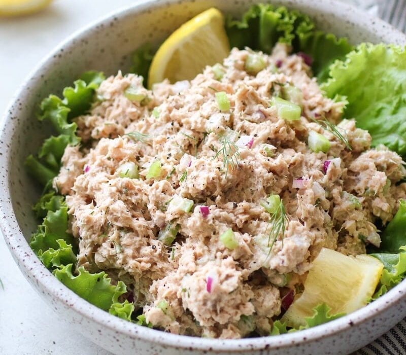 A speckled white bowl holding fresh salmon salad that is laying on top of healthy green leafy lettuce with a couple of lemon wedges.