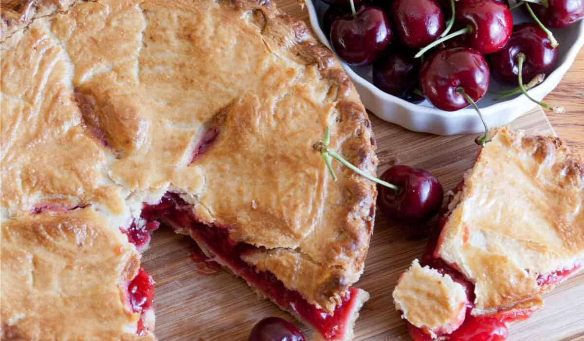 Overhead image of a cherry pie with a slice taken out of it on a wooden cutting board