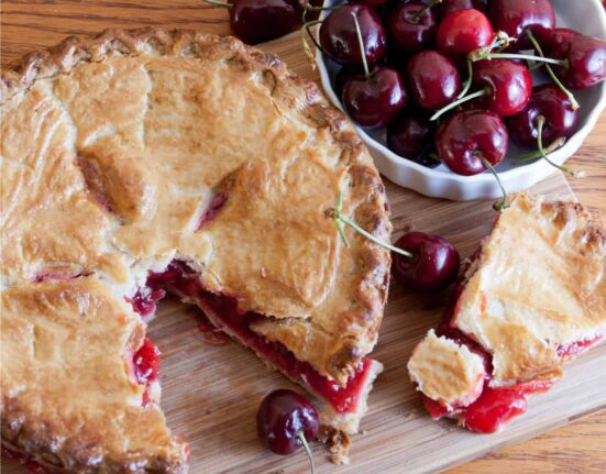 Overhead image of a cherry pie with a slice taken out of it on a wooden cutting board