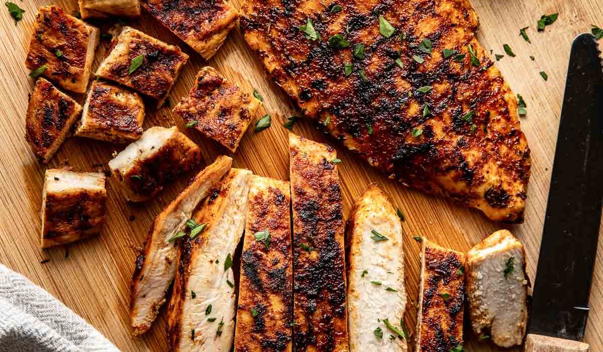 Overhead view of a wooden cutting board with blackened chicken breast on it, one sliced and one whole with lime on the side.