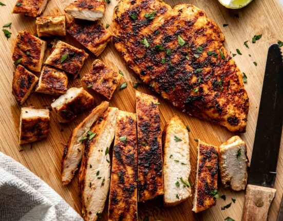 Overhead view of a wooden cutting board with blackened chicken breast on it, one sliced and one whole with lime on the side.