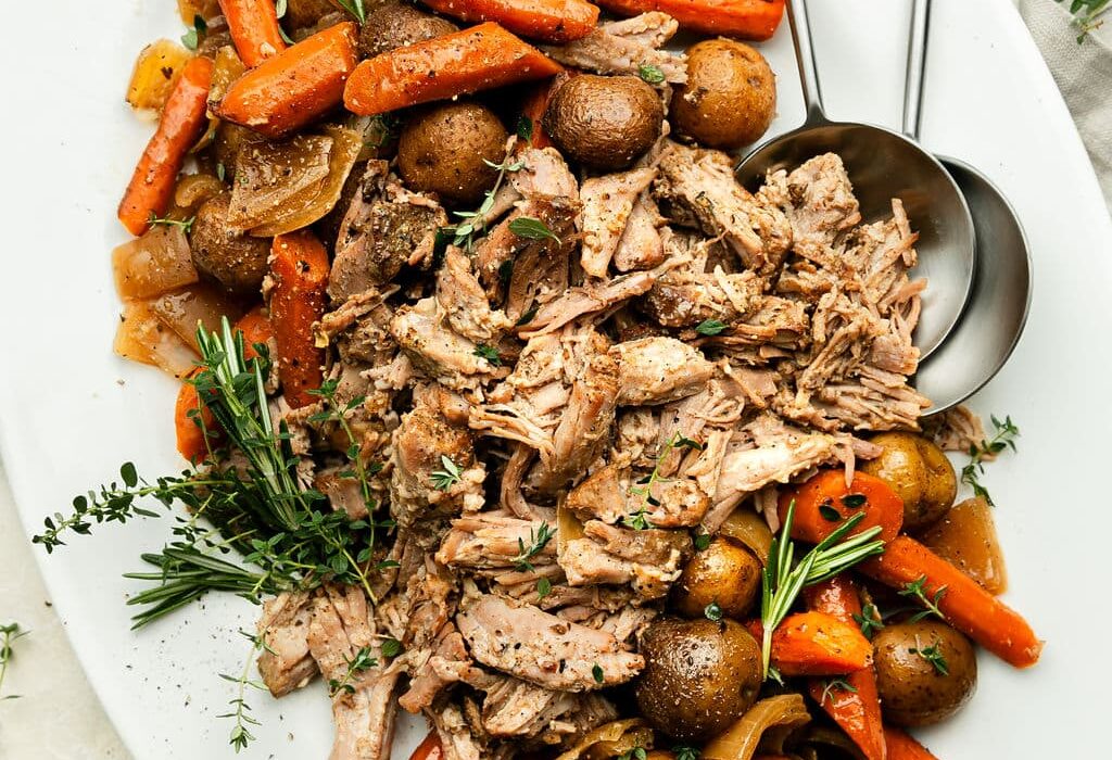 An overhead view of a platter of Crock Pot Pork Roast with onions, carrots, and potatoes. The dish is garnished with fresh thyme and rosemary.
