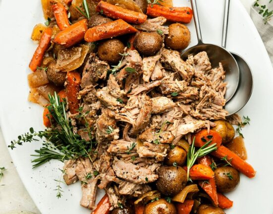 An overhead view of a platter of Crock Pot Pork Roast with onions, carrots, and potatoes. The dish is garnished with fresh thyme and rosemary.