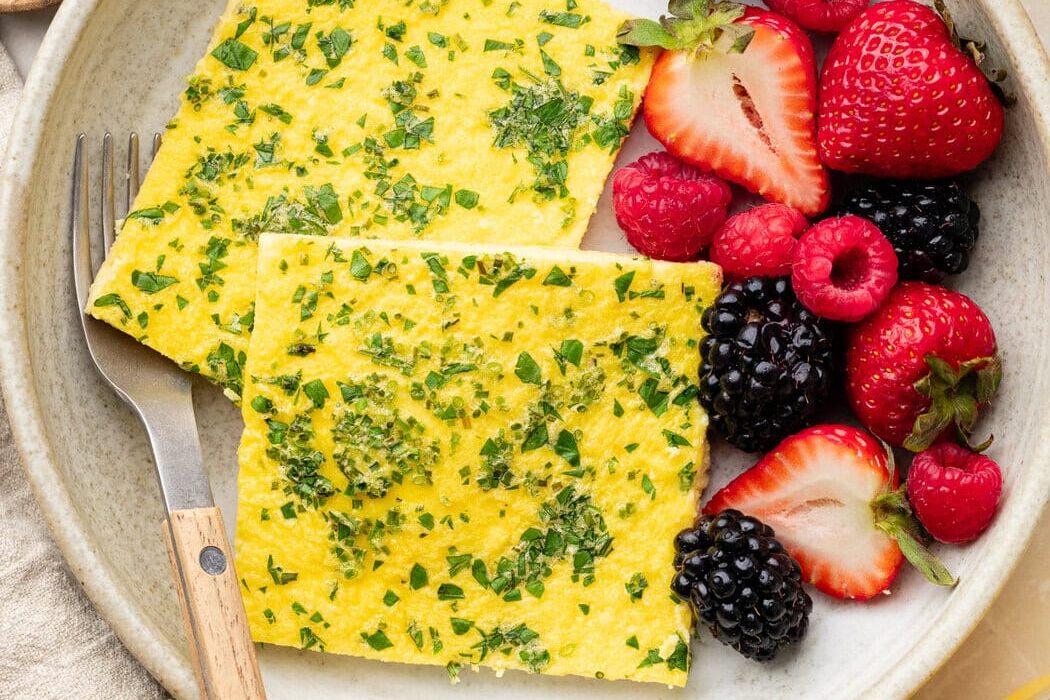 Overhead view of a white ceramic plate filled with 2 squares of sheet pan eggs topped with fresh herbs and colorful fruit on the side.