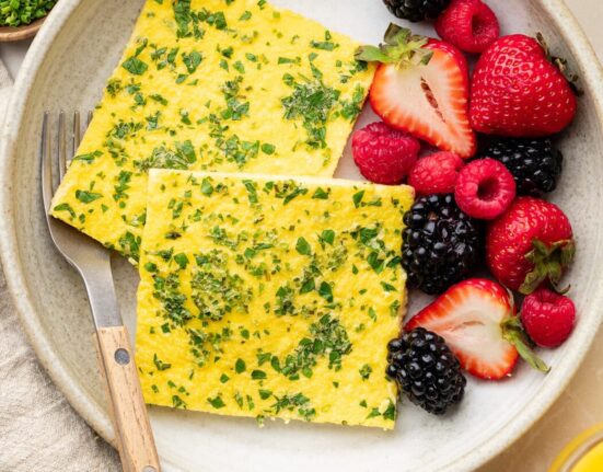 Overhead view of a white ceramic plate filled with 2 squares of sheet pan eggs topped with fresh herbs and colorful fruit on the side.