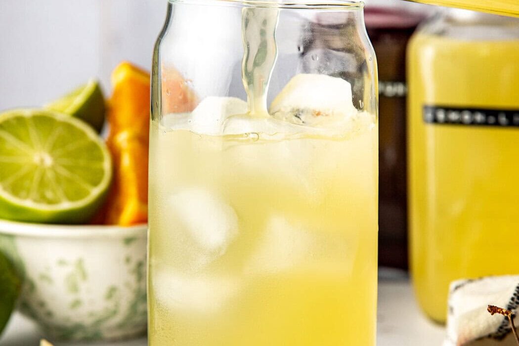 Close up view of a hand pouring a yellow Homemade Gatorade mixture into a glass of ice with fresh fruit in the background.