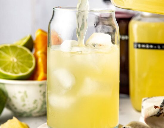 Close up view of a hand pouring a yellow Homemade Gatorade mixture into a glass of ice with fresh fruit in the background.