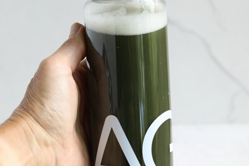 Close up view of a hand holding a clear bottle of mixed AG1 greens showing the green color and foam on top.