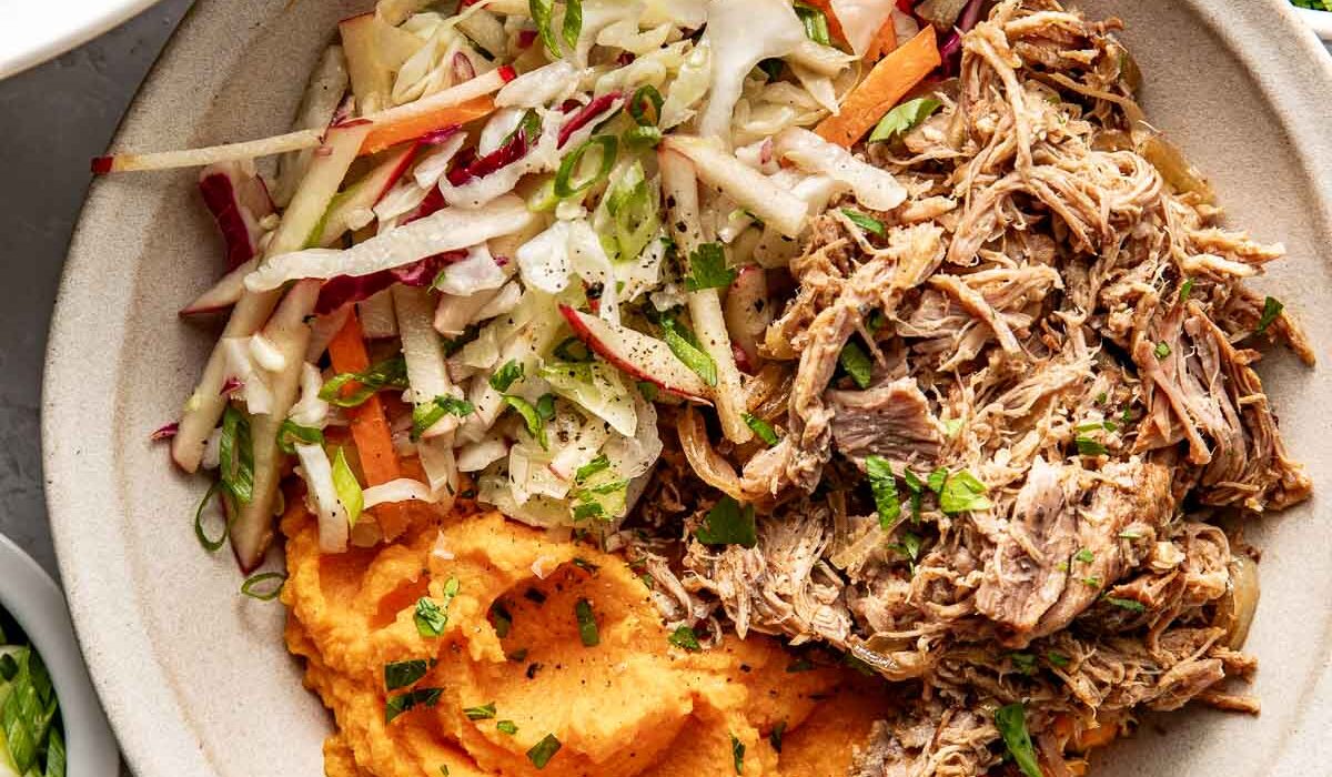 Overhead view of a white bowl filled with deep orange mashed sweet potatoes, pulled pork, and colorful fresh apple slaw.