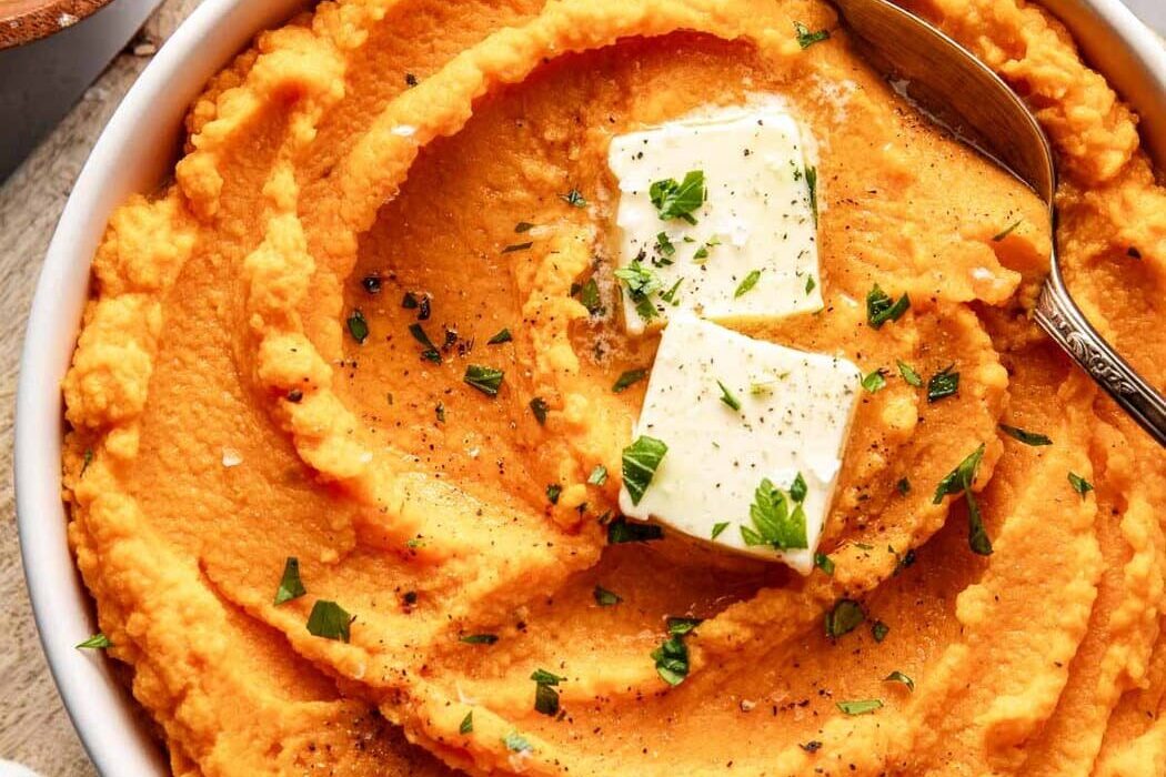 Overhead view of a bowl of vibrant orange Mashed Sweet Potatoes topped with butter and fresh herbs.