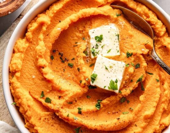 Overhead view of a bowl of vibrant orange Mashed Sweet Potatoes topped with butter and fresh herbs.