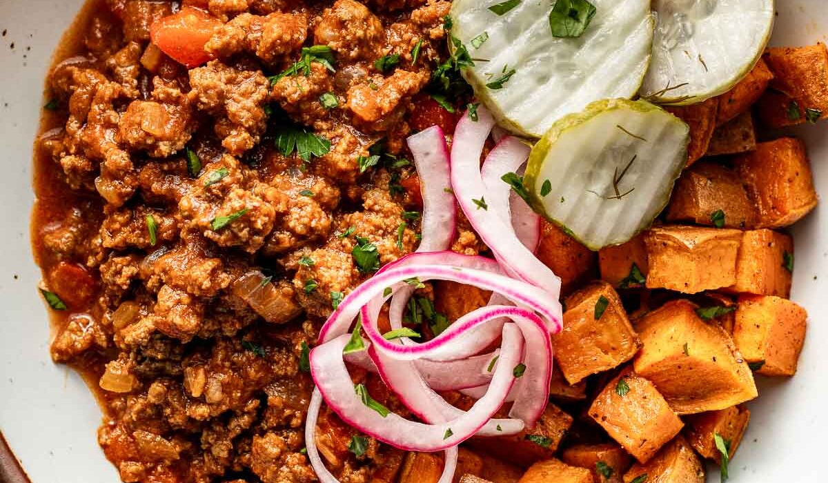 Overhead view of a bowl of sloppy joe meat, roasted sweet potatoes, pickled red onions, and crisp pickle chips.