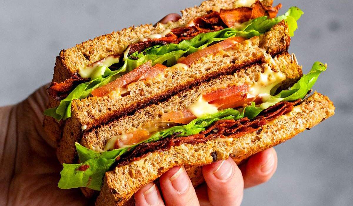 A hand holding a vegan BLT sandwich with carrot bacon.