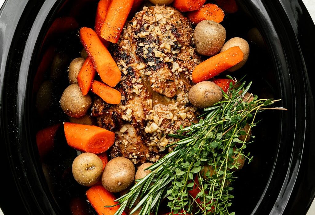 Overhead view of vegetables and a seasoned covered roast in a black crockpot garnished with a bundle of fresh herbs.
