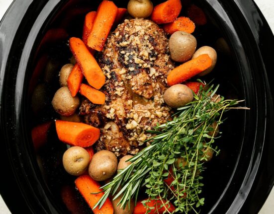 Overhead view of vegetables and a seasoned covered roast in a black crockpot garnished with a bundle of fresh herbs.