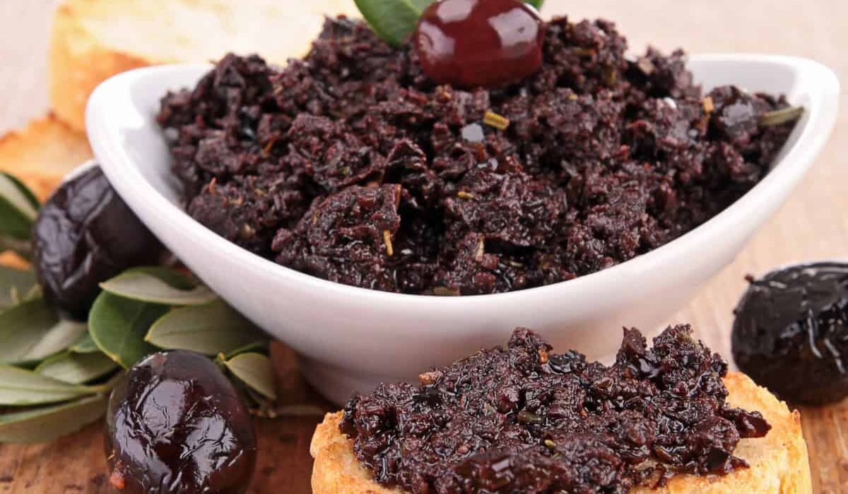 Olive Tapenade in a white bowl and spread on a piece of bread