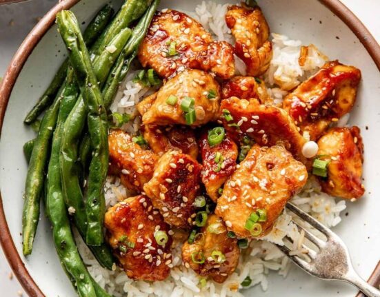 Overhead view of a plate of Sesame Chicken on top of white rice sprinkled with green onions next to roasted green beans.