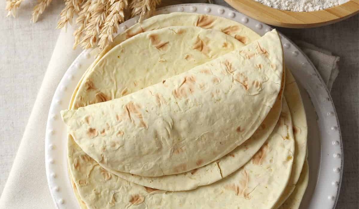Overhead image of Flour Tortillas stacked on a white plate next to a bowl of flour