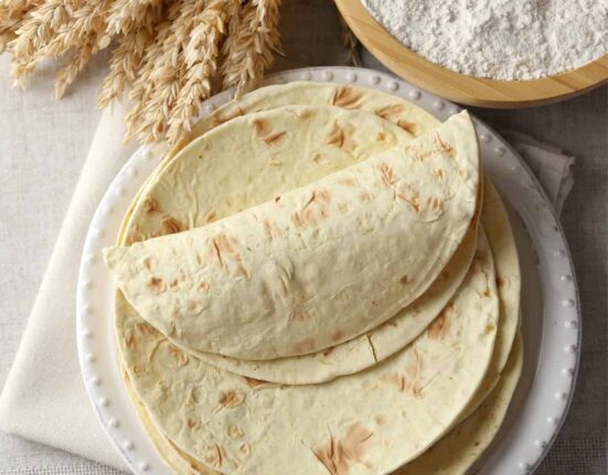 Overhead image of Flour Tortillas stacked on a white plate next to a bowl of flour