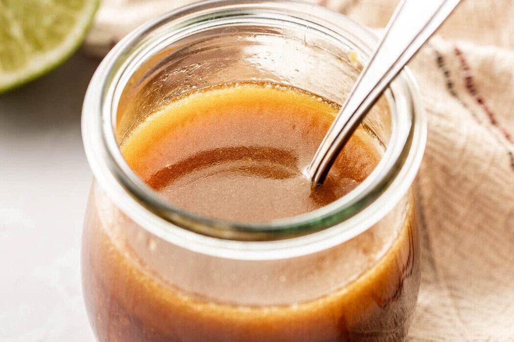 Close up view of a glass jar with a spoon in it filled with Sesame Ginger Dressing with Lime in the background.