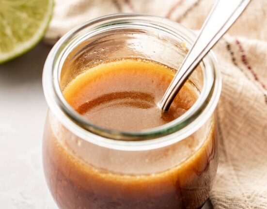 Close up view of a glass jar with a spoon in it filled with Sesame Ginger Dressing with Lime in the background.