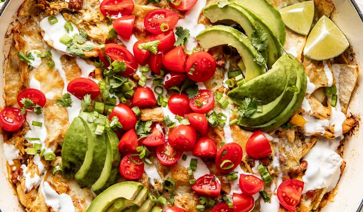Overhead view of a skillet filled with chicken enchiladas topped with sour cream, avocado, and tomatoes.