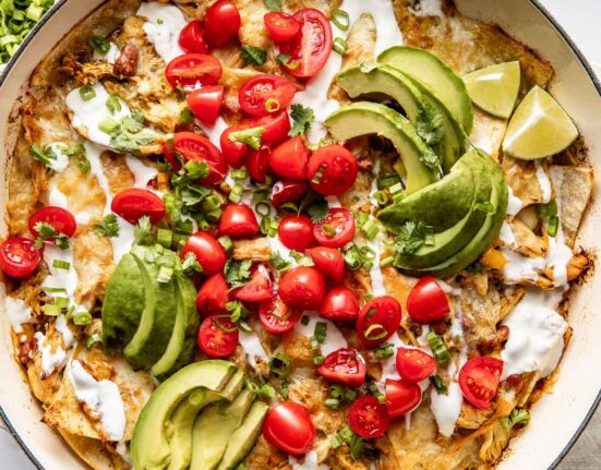 Overhead view of a skillet filled with chicken enchiladas topped with sour cream, avocado, and tomatoes.