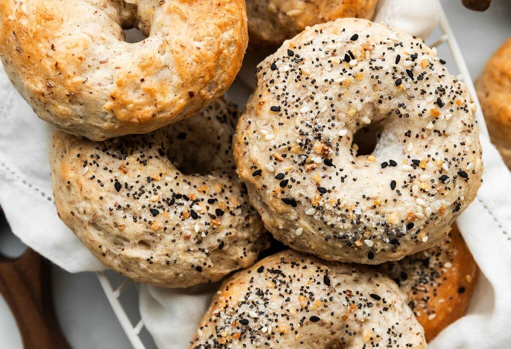Freshly baked Greek yogurt bagels in a wire basket. Some of the bagels are top with Everything Bagel Seasoning.