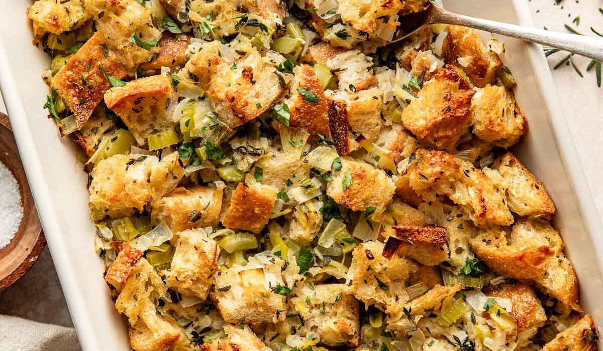 Overhead view of a white baking dish filled with stuffing topped with fresh herbs and black pepper.