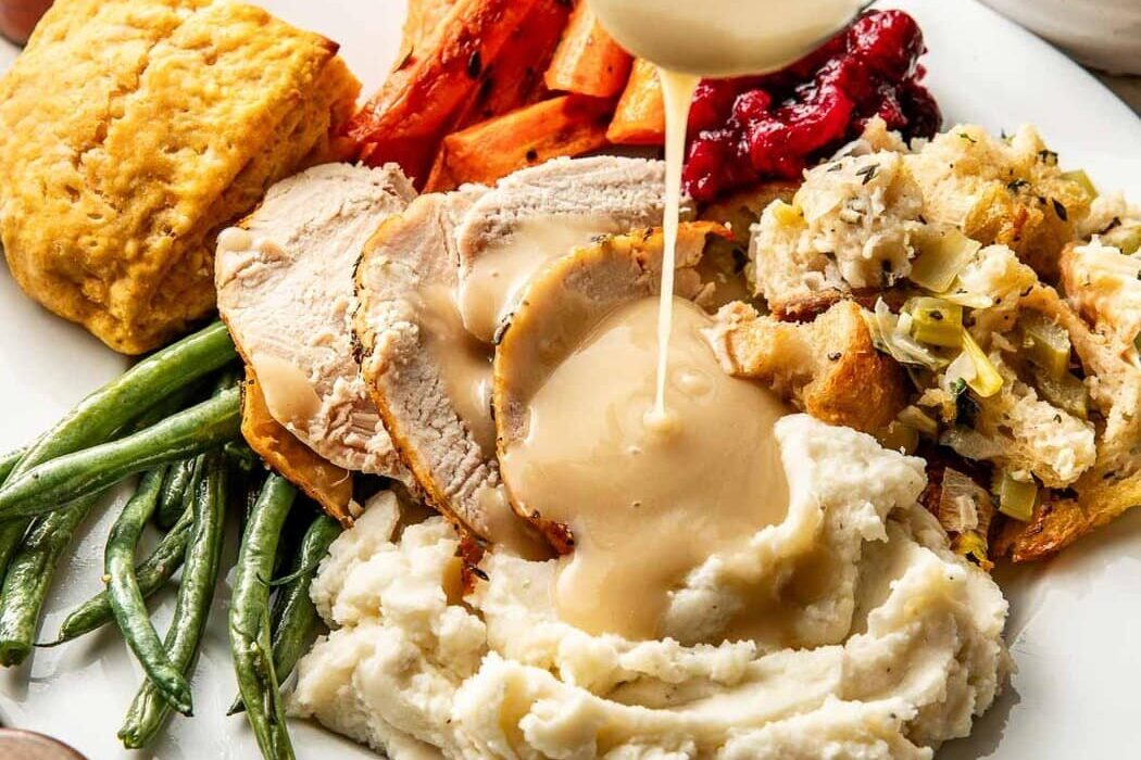 Close up view of a white plate with roasted vegetables, stuffing, a sweet potato biscuit, and sliced turkey on top of mashed potatoes with gravy pouring over it.