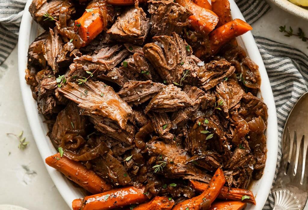 Close up view of an oval platter filled with shredded venison roast surrounded by carrots and topped with a brown gravy and sprinkled with fresh herbs and black pepper.
