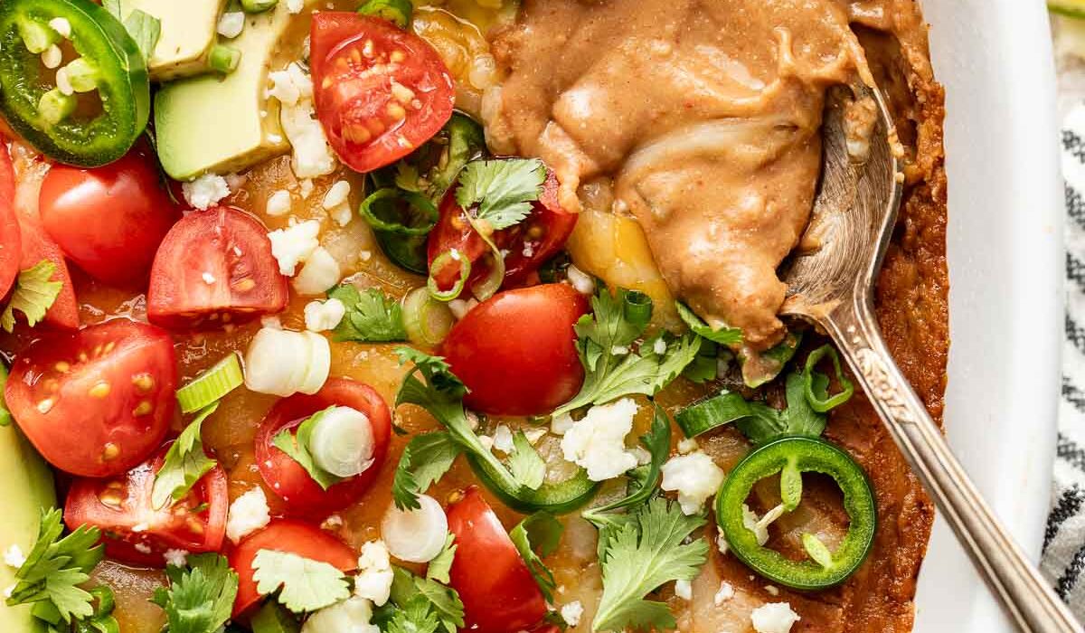 Overhead view of a white baking dish filled with Homemade Bean Dip and topped with fresh cilantro, tomatoes, and sliced jalapeno with a spoon in the side.