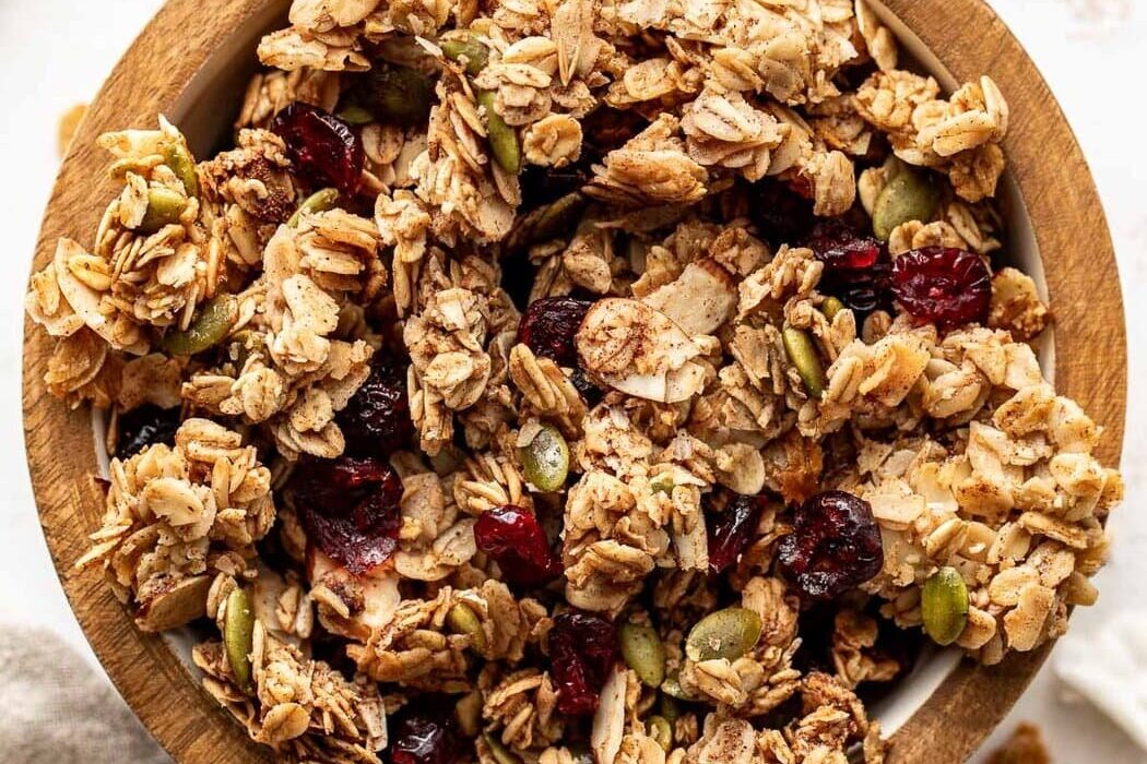 Overhead view of a small wooden bowl filled with Homemade Granola showing the crunchy texture and dried fruit pieces throughout the mixture.