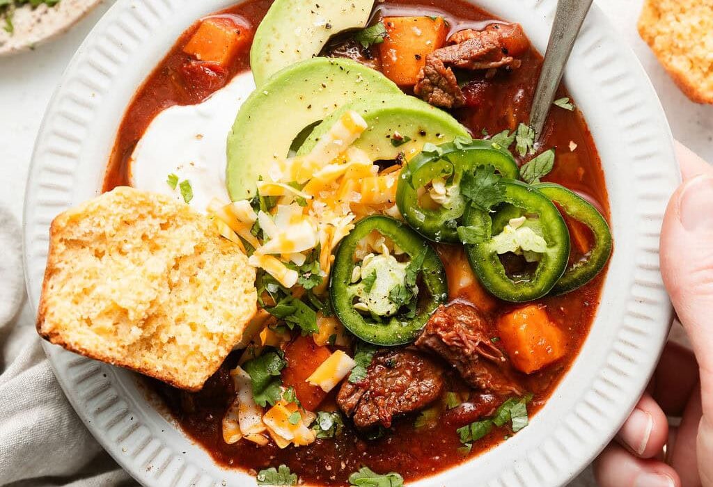 Overhead view of a bowl of Slow Cooker Chuck Roast Chili With Sweet Potatoes and a corn bread muffin on the side.
