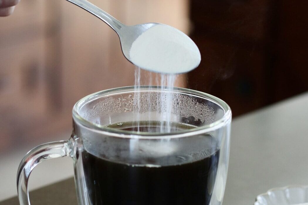 Close up view of a hand pouring a spoonful of collagen into a glass mug of coffee.