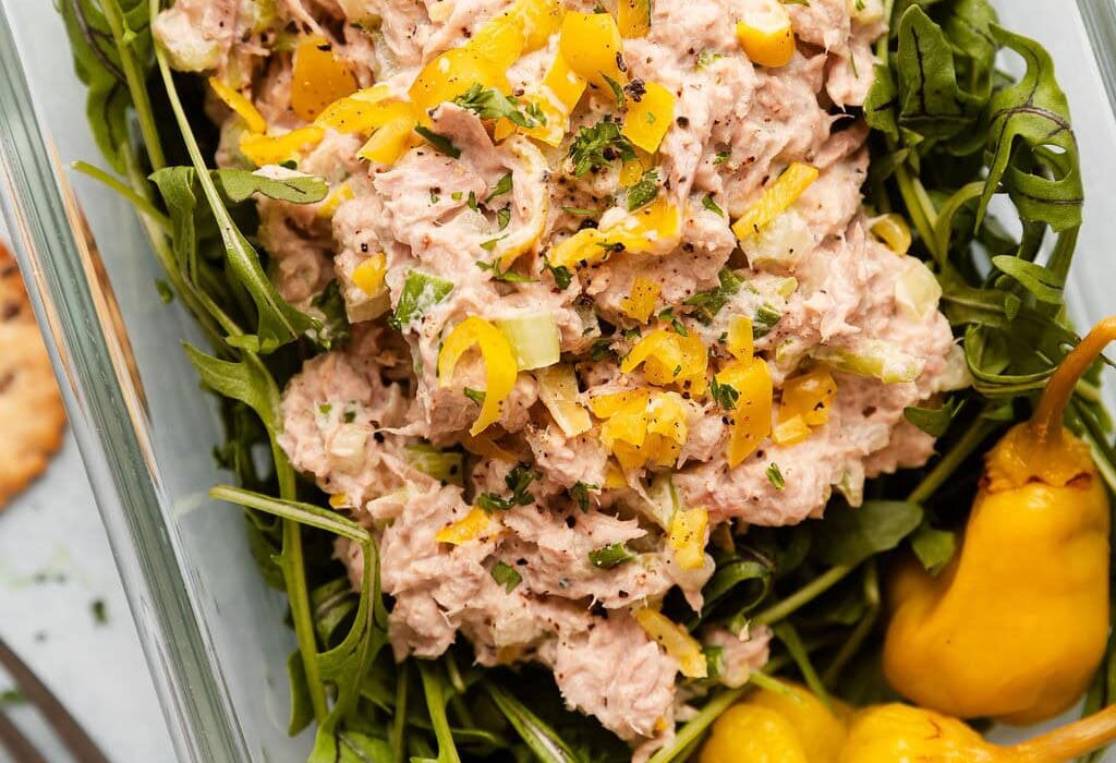 Overhead view of a glass meal prep container filled with arugula and topped with Tuna Salad garnished with pickled peppers.