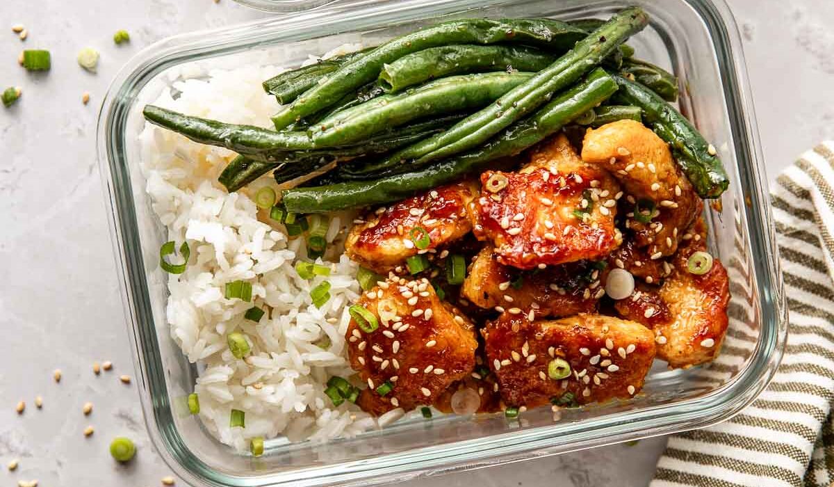 Overhead view of three glass meal prep containers filled with sesame chicken, white rice, and green beans sprinkled with sesame seeds.