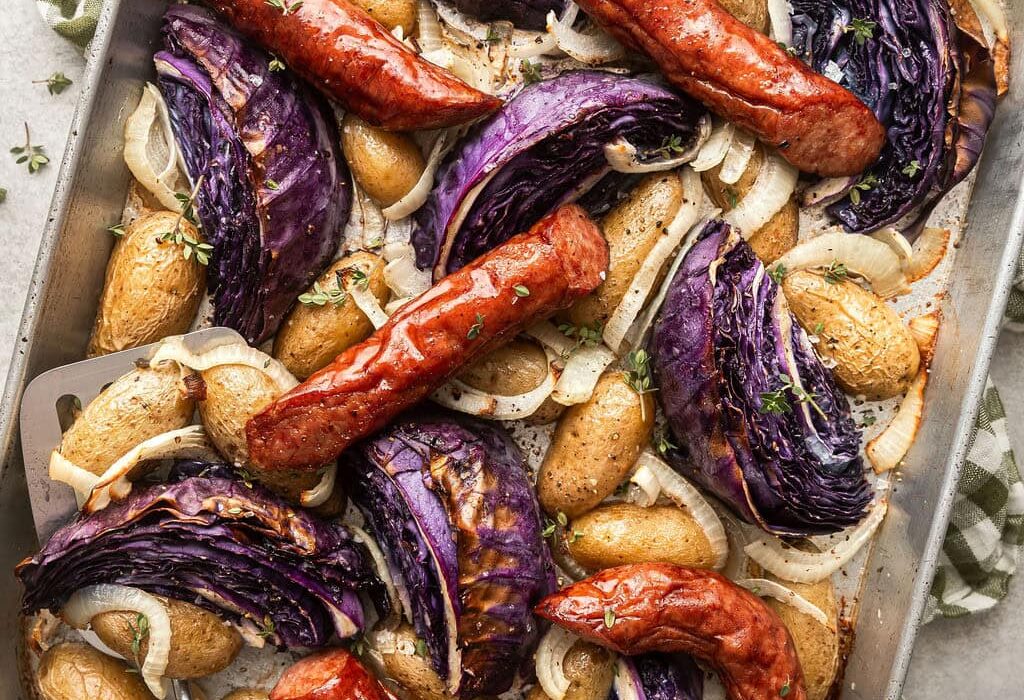 Overhead view of a sheet pan of kielbasa and potatoes with wedges of purple cabbage topped with fresh herbs and black pepper.