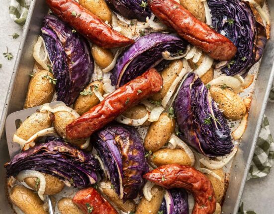 Overhead view of a sheet pan of kielbasa and potatoes with wedges of purple cabbage topped with fresh herbs and black pepper.