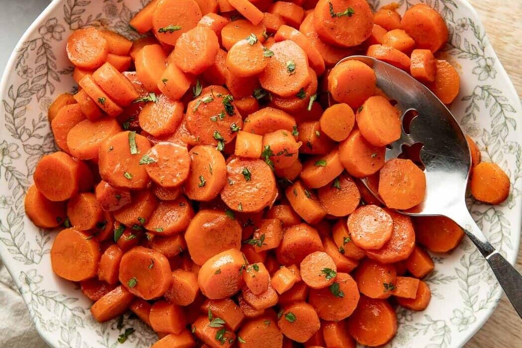 Overhead view of a decorative bowl filled with sliced cooked carrots topped with fresh herbs and a serving spoon in the side of the bowl.