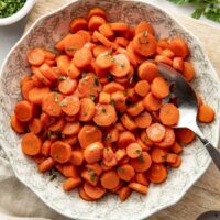 Overhead view of a decorative bowl filled with sliced cooked carrots topped with fresh herbs and a serving spoon in the side of the bowl.