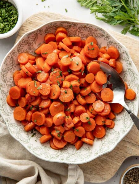 Overhead view of a decorative bowl filled with sliced cooked carrots topped with fresh herbs and a serving spoon in the side of the bowl.