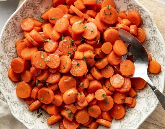 Overhead view of a decorative bowl filled with sliced cooked carrots topped with fresh herbs and a serving spoon in the side of the bowl.