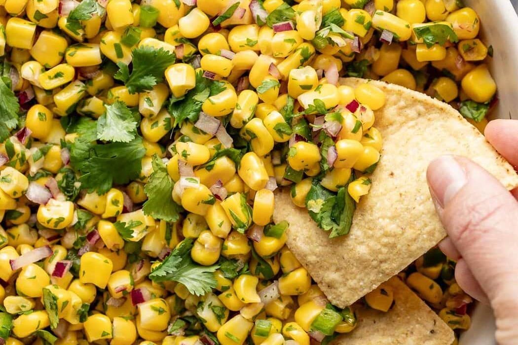 Overhead view of a bowl of Copycat Chipotle Corn Salsa with a hand scooping a tortilla chip into the mixture.