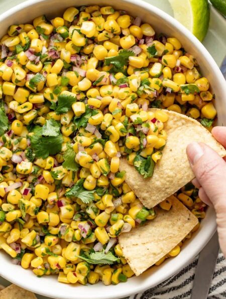Overhead view of a bowl of Copycat Chipotle Corn Salsa with a hand scooping a tortilla chip into the mixture.