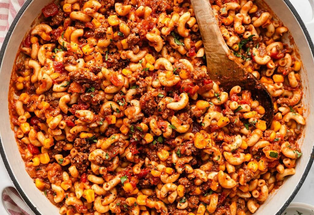 Overhead view of a skillet filled with goulash topped with fresh herbs and a wooden spoon stuck in the side.