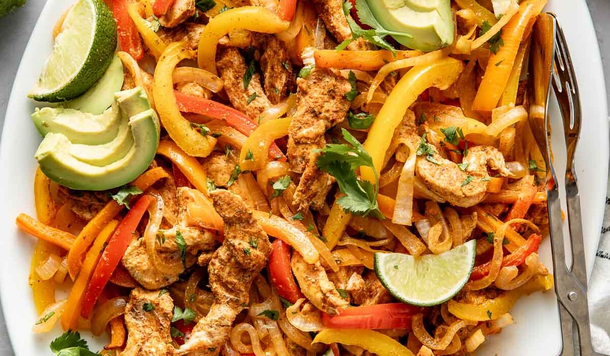 Overhead view of a platter filled with Chicken Fajitas and garnished with avocado, limes, and cilantro.