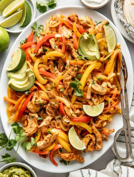 Overhead view of a platter filled with Chicken Fajitas and garnished with avocado, limes, and cilantro.