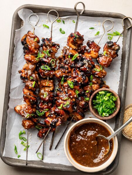 Overhead view of a sheet pan of Korean BBQ chicken skewers topped with green onions and sesame seeds.
